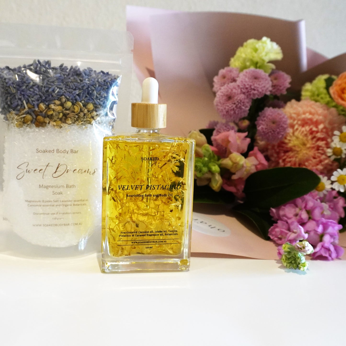 Bottle of 'Velvet Petal' oil with a bouquet of flowers and a bag labeled 'Sweet Dreams' on a white surface.