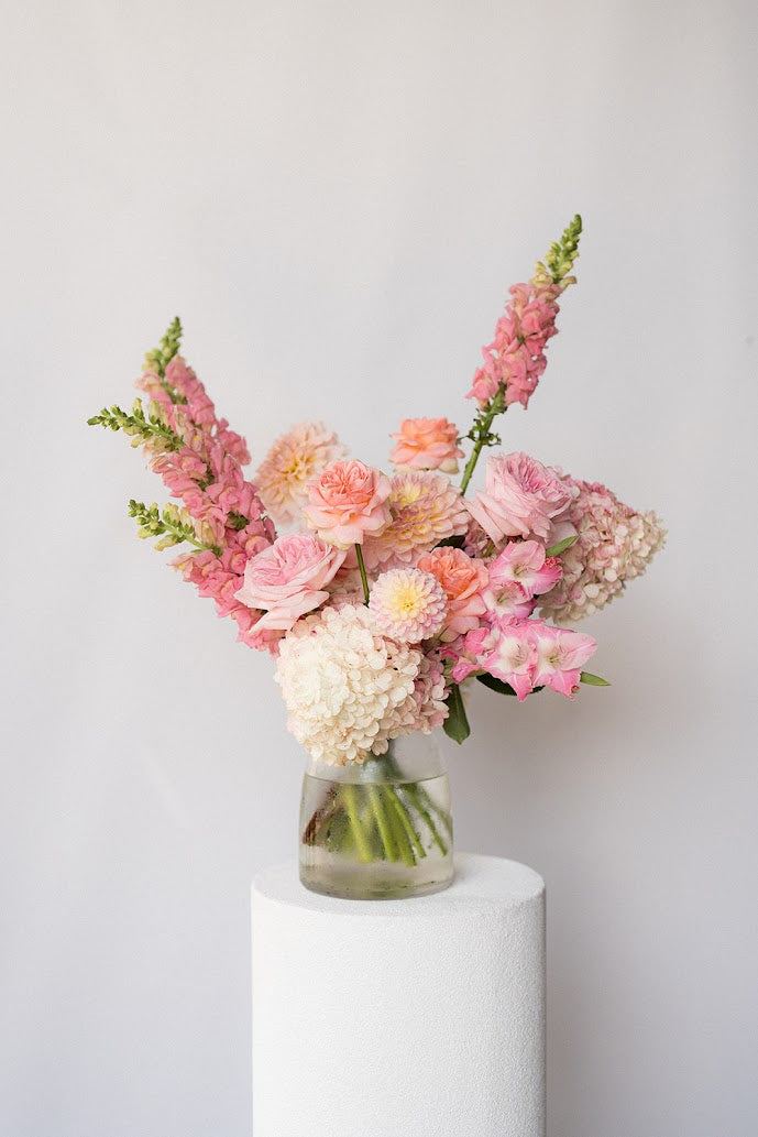Bouquet of pink and white flowers in a clear vase on a white pedestal against a light gray background