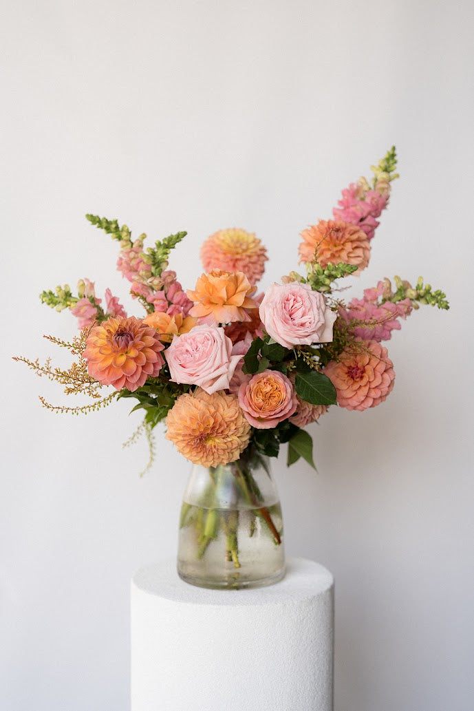 Bouquet of pink and orange flowers in a clear vase on a white pedestal against a light gray background