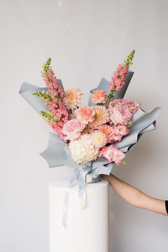 Bouquet of pink and white flowers wrapped in gray paper with a hand holding it against a light gray background.