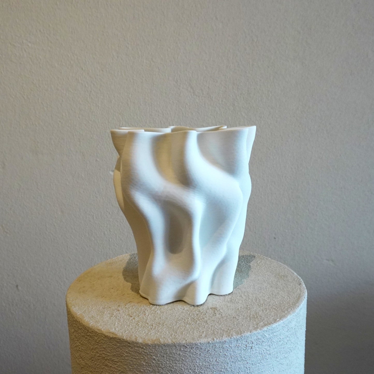White abstract ceramic vase on a cylindrical stone pedestal against a plain wall.
