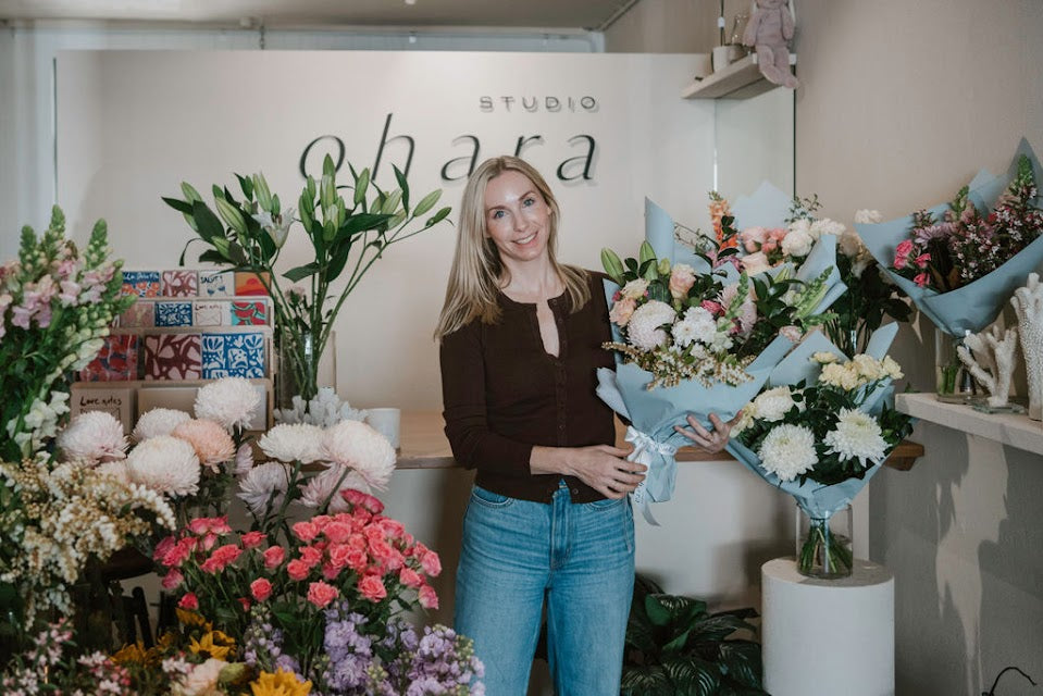 Woman holding a bouquet of flowers in a flower shop with a 'Studio Ohara' sign in the background.