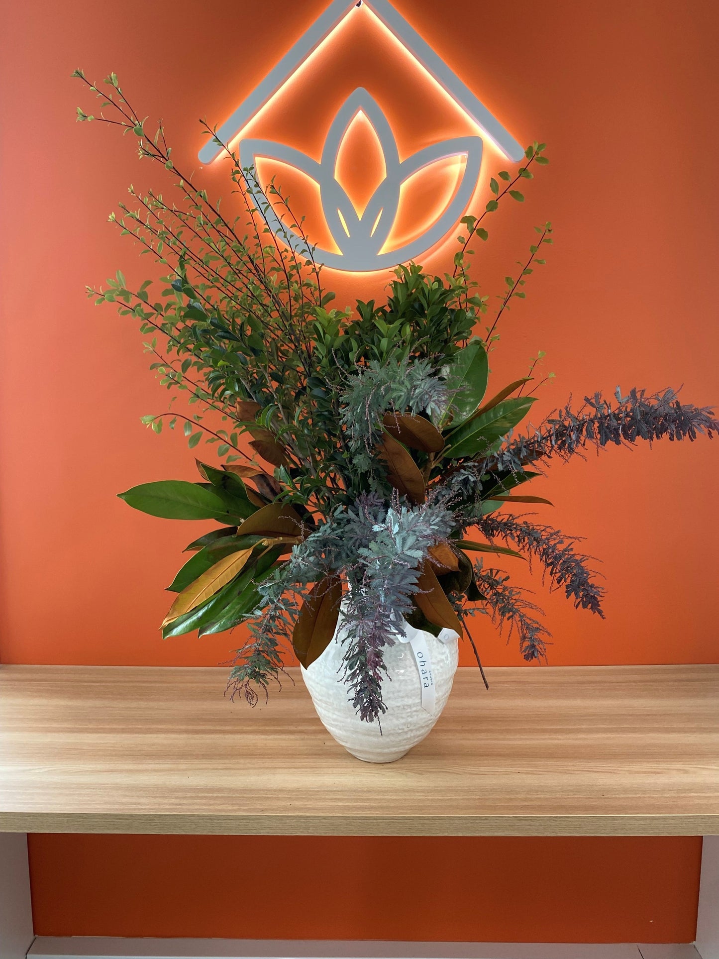 Floral arrangement in a vase on a wooden surface with an orange wall and logo in the background