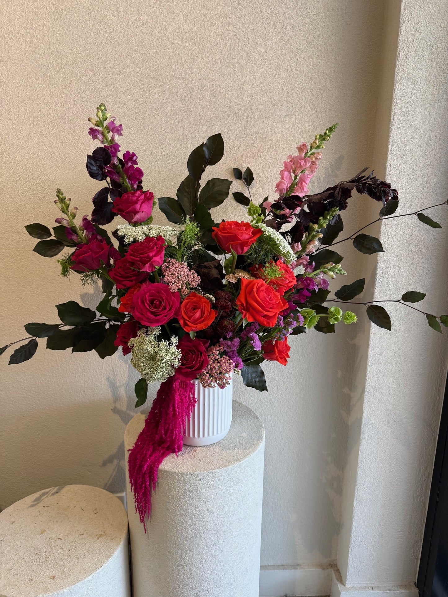 Luxe christmas style arrangement with red and pink roses, purple stock flowers and burgundy foliage in white vase on beige pedestal