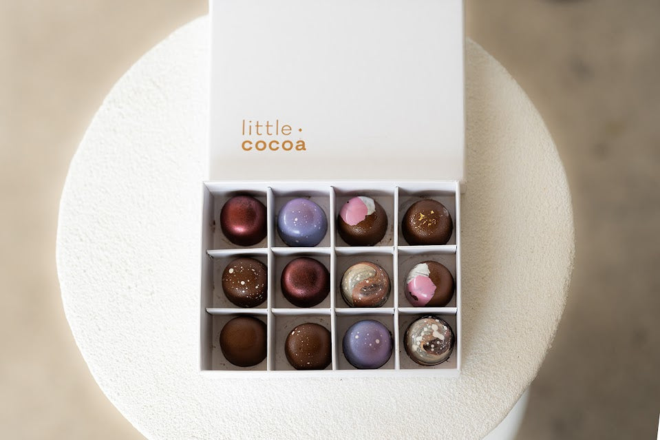 Box of assorted chocolates with 'little cocoa' branding on a white stand.