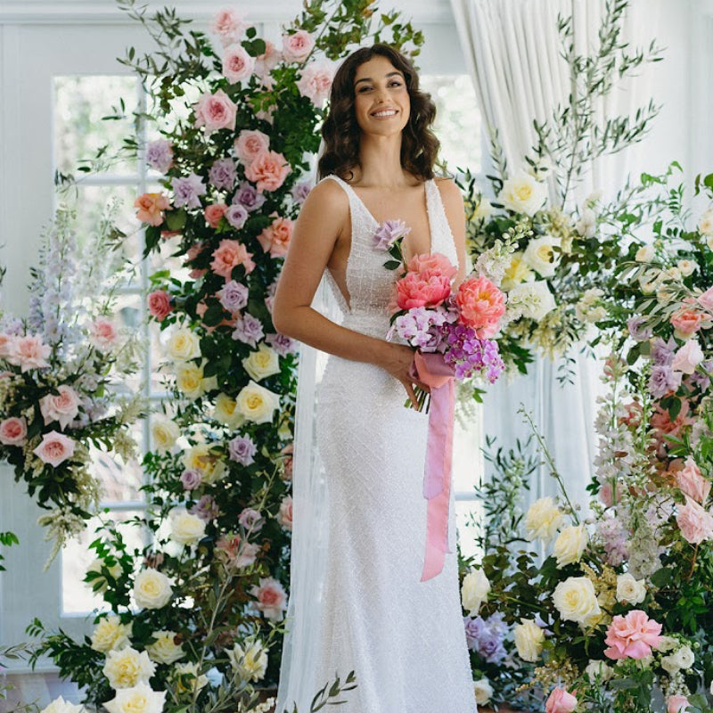 Woman in a white wedding dress holding flowers amidst floral decorations in a bright room.
